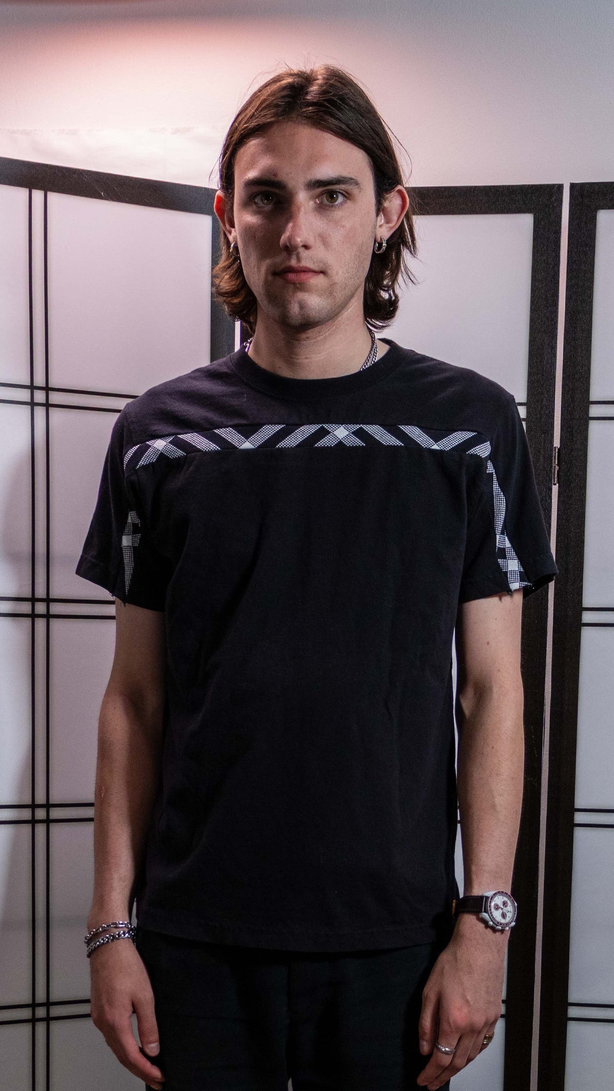 A man wearing a black short-sleeve t-shirt with white geometric patterns on the sleeve, standing in front of a white wall with black grid lines.