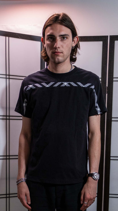 A man wearing a black short-sleeve t-shirt with white geometric patterns on the sleeve, standing in front of a white wall with black grid lines.