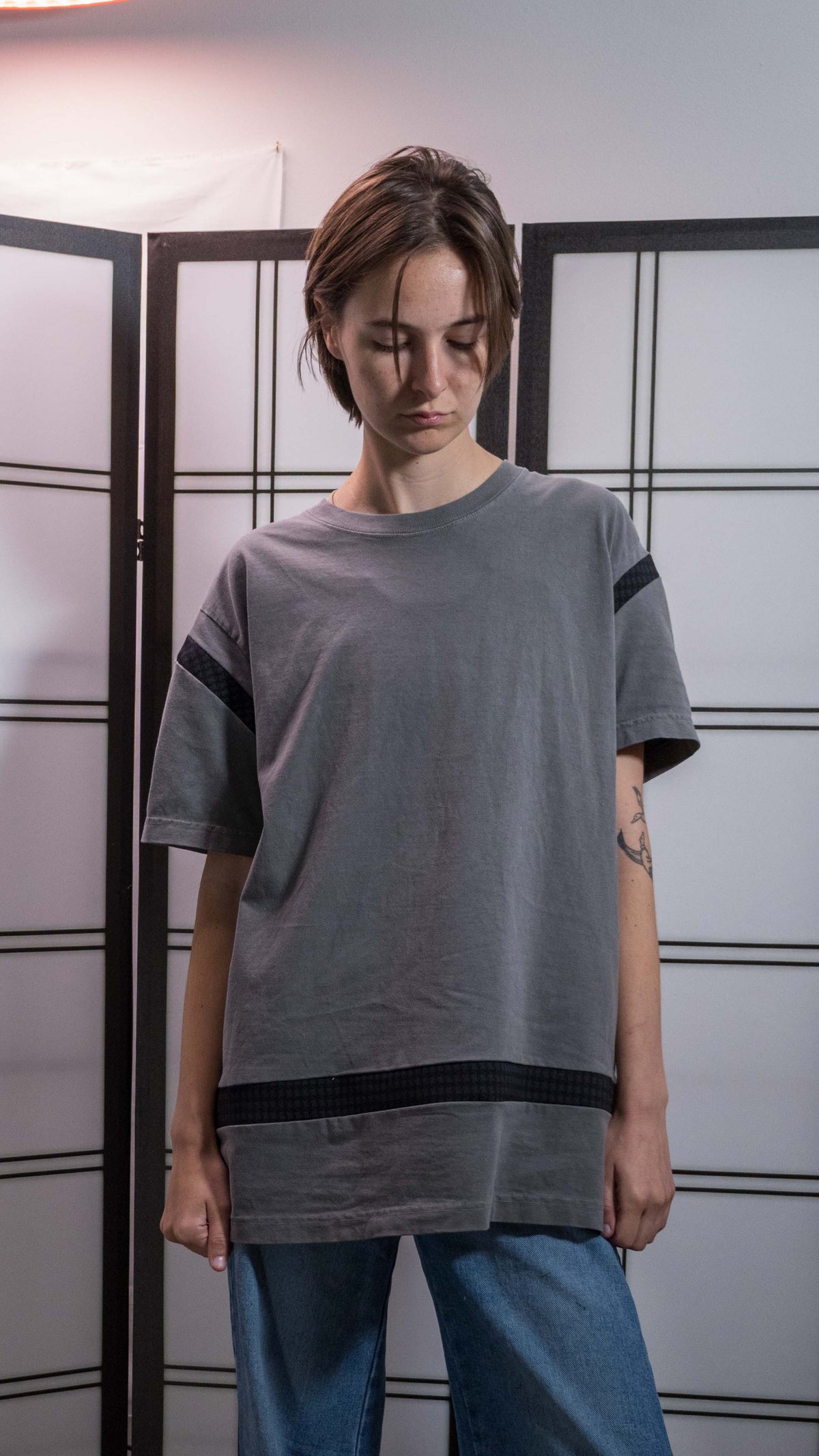 A person wearing a gray oversized short-sleeve t-shirt with black stripe details on the sleeves and hem, paired with blue jeans, standing in front of a white shoji screen.