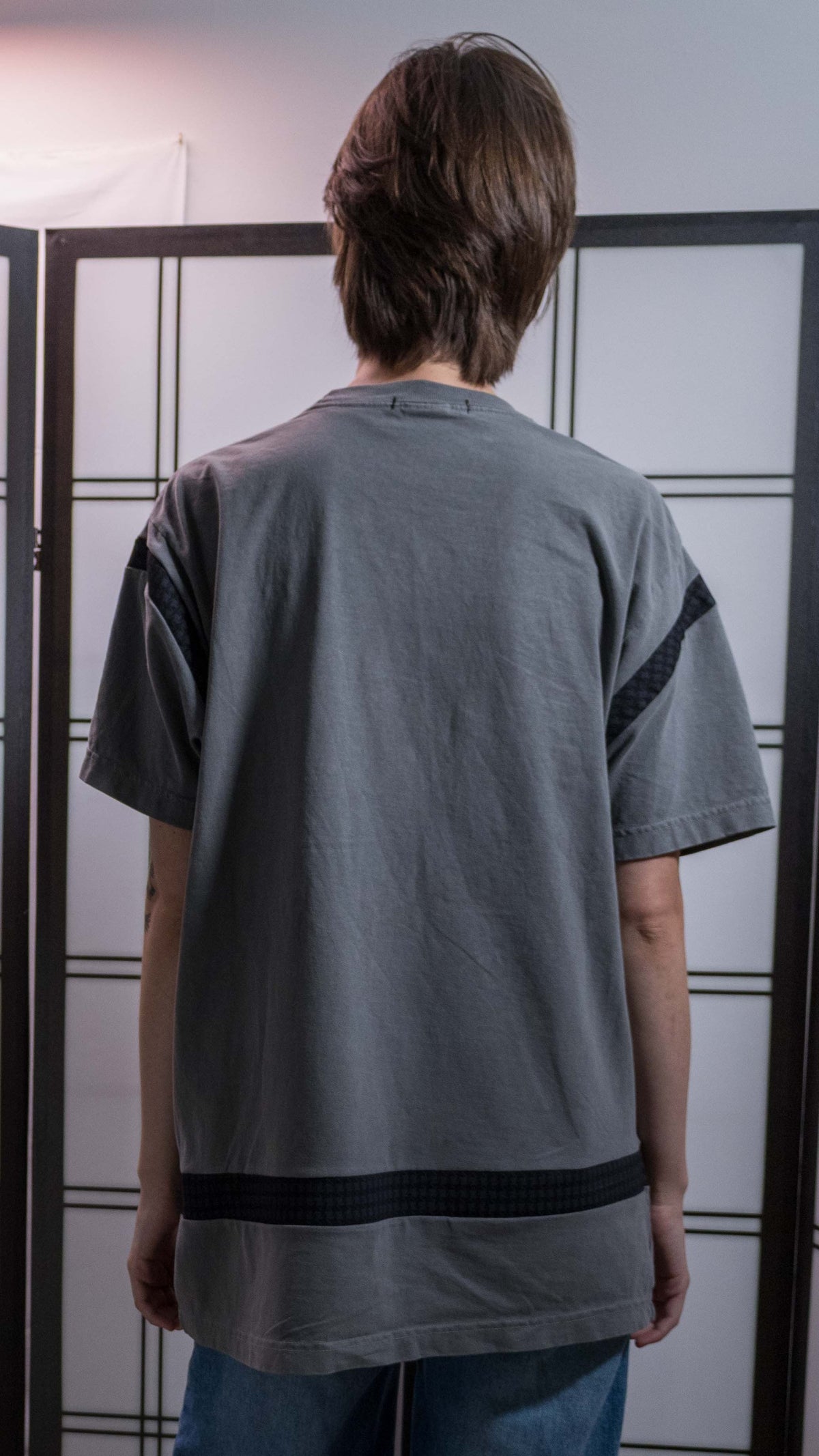 A person viewed from behind wearing a loose gray t-shirt with black stripes on the sides and blue jeans, standing in front of a white shoji screen.